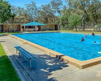 A clear outdoor swimming pool surrounded by trees, with people enjoying the water on a sunny day. Benches line the pool's edge.