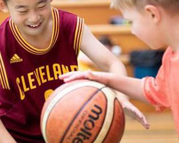 Two children play basketball, smiling and reaching for the ball.