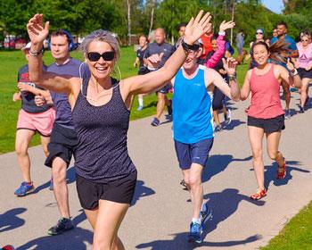 Image of people running on a footpath. The woman at the front of the group is smiling with her hands in the air