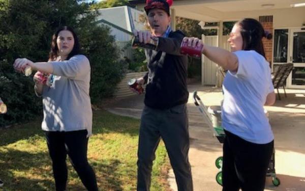 Three people engage in a playful activity outdoors, holding bottles while standing in a grassy area, with a building in the background.