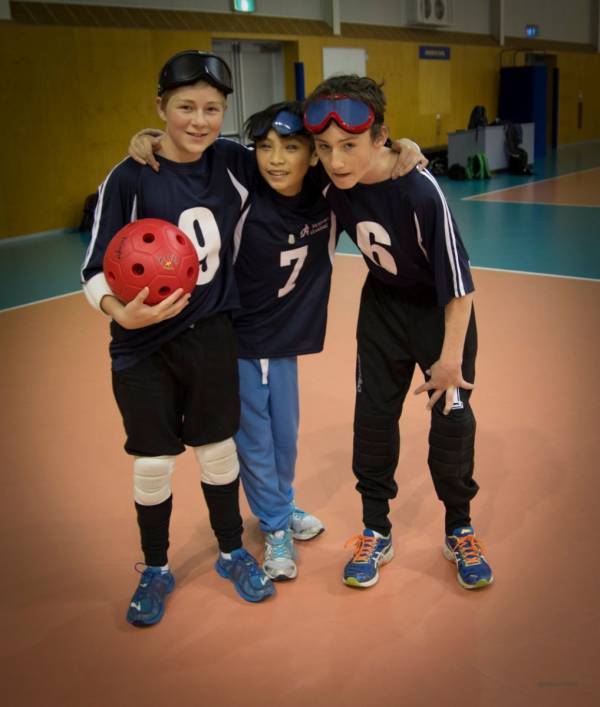 Pictured are three children stanging together holding a goalball and wearing goalball goggles 